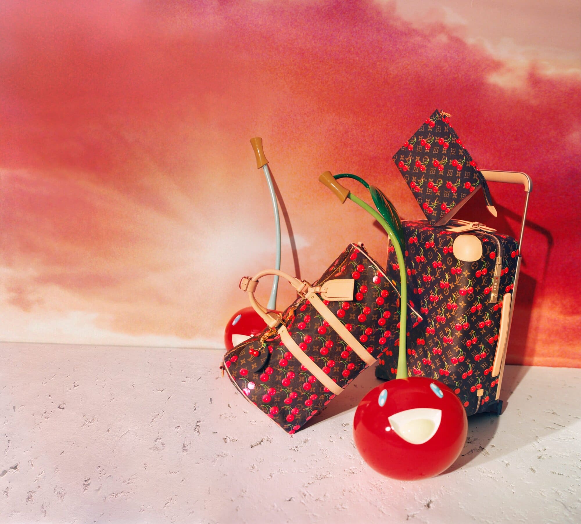 Louis Vuitton x Murakami cherry print luggage and handbags with stylized smiling cherry sculptures against a vibrant sunset background.
