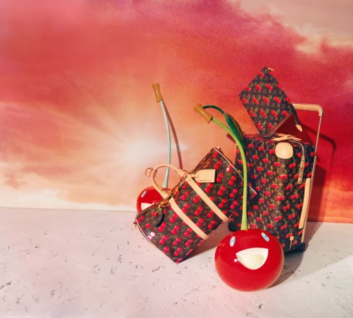 Louis Vuitton x Murakami cherry print luggage and handbags with stylized smiling cherry sculptures against a vibrant sunset background.