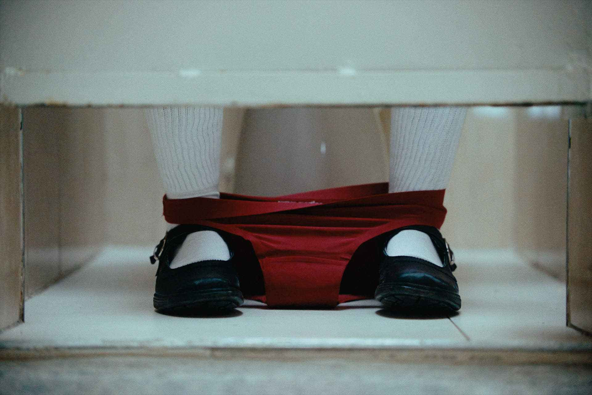 Close-up of a person seated on a toilet wearing red underwear down around their ankles, white socks, and black shoes.