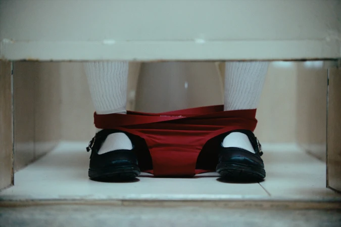 Close-up of a person seated on a toilet wearing red underwear down around their ankles, white socks, and black shoes.