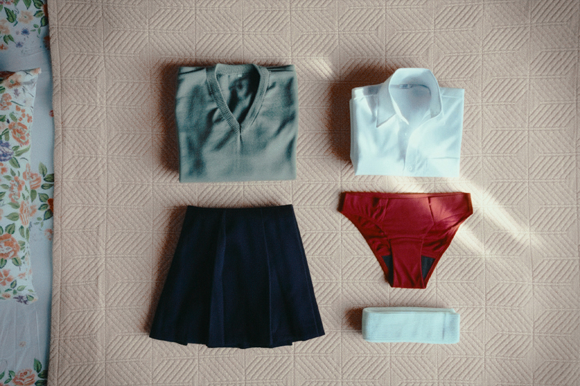 Flat lay of neatly folded school uniform pieces including a grey sweater, white shirt, dark skirt, and red undergarments.