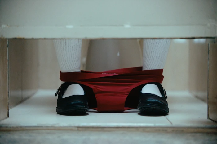 Close-up of legs wearing white socks and black Mary Jane shoes with red underwear bunched around the ankles inside a stall