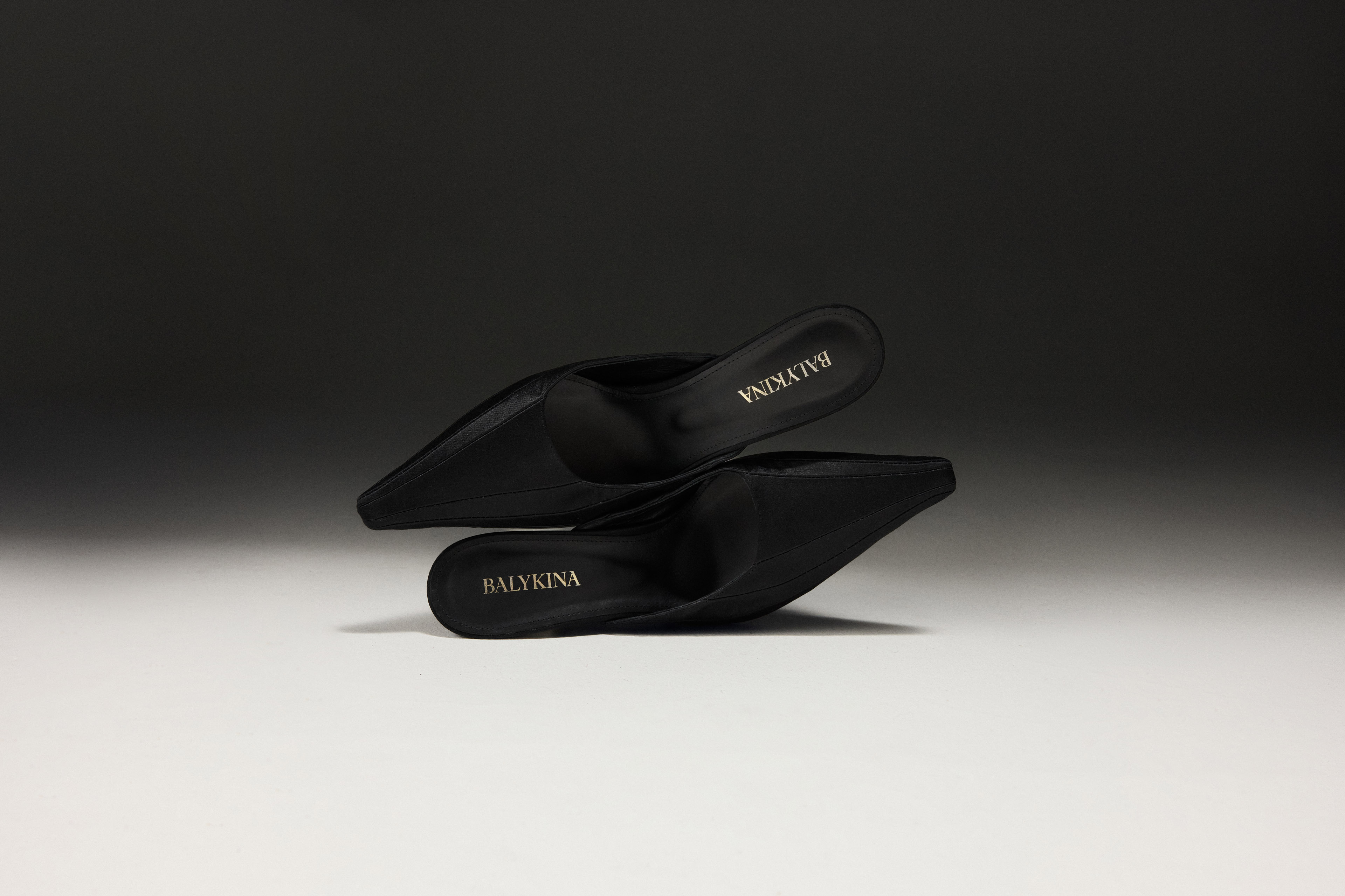 Two black high-heeled mules with pointed toes stacked horizontally against a gradient grey studio background