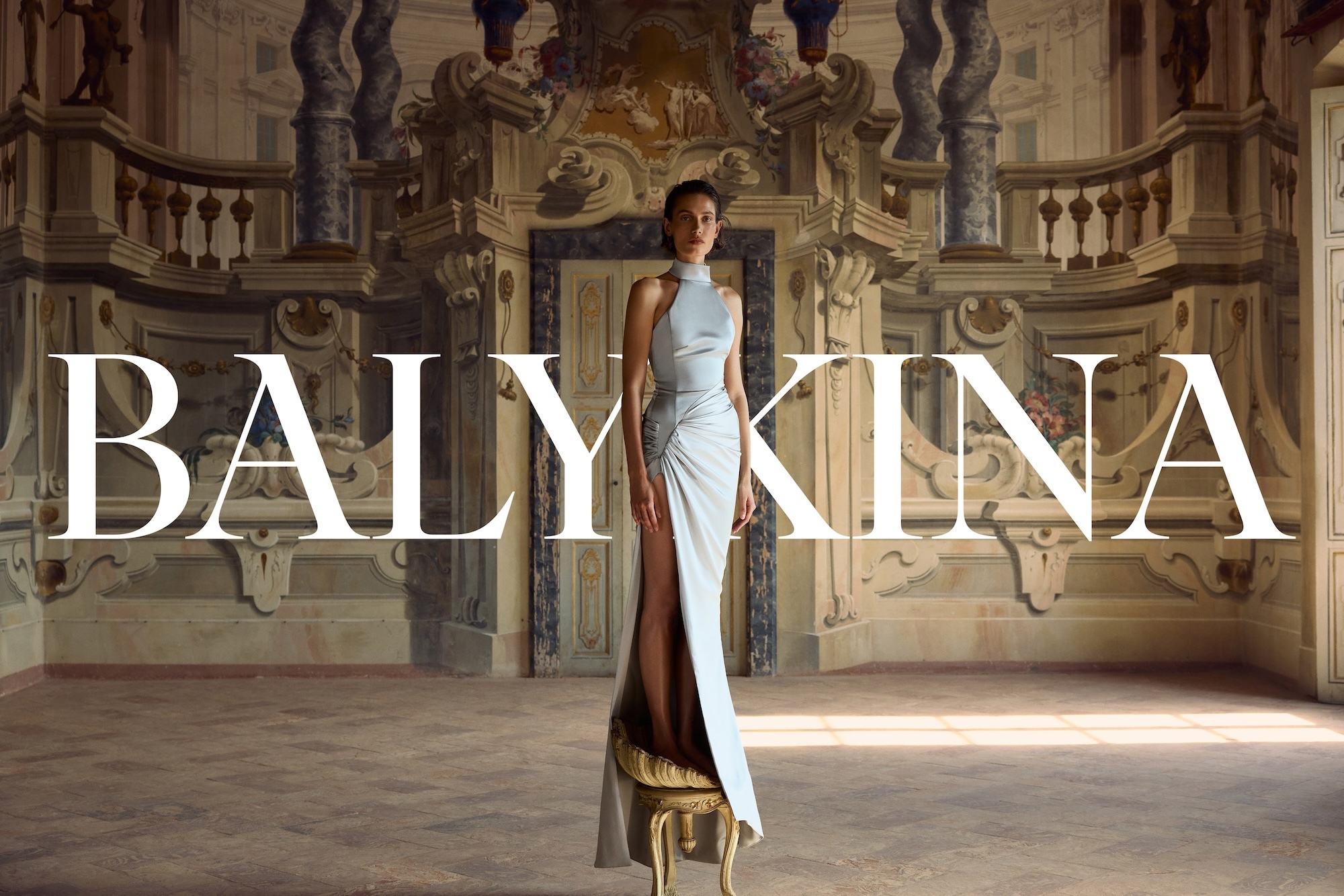 Model standing on a small stool in a silk halter dress inside an ornate hall with arched walls and classical architectural details.