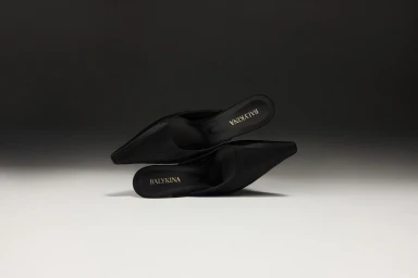 Two black high-heeled mules with pointed toes stacked horizontally against a gradient grey studio background