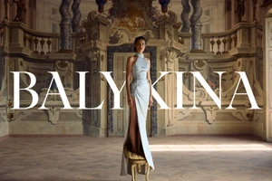 Model standing on a small stool in a silk halter dress inside an ornate hall with arched walls and classical architectural details.
