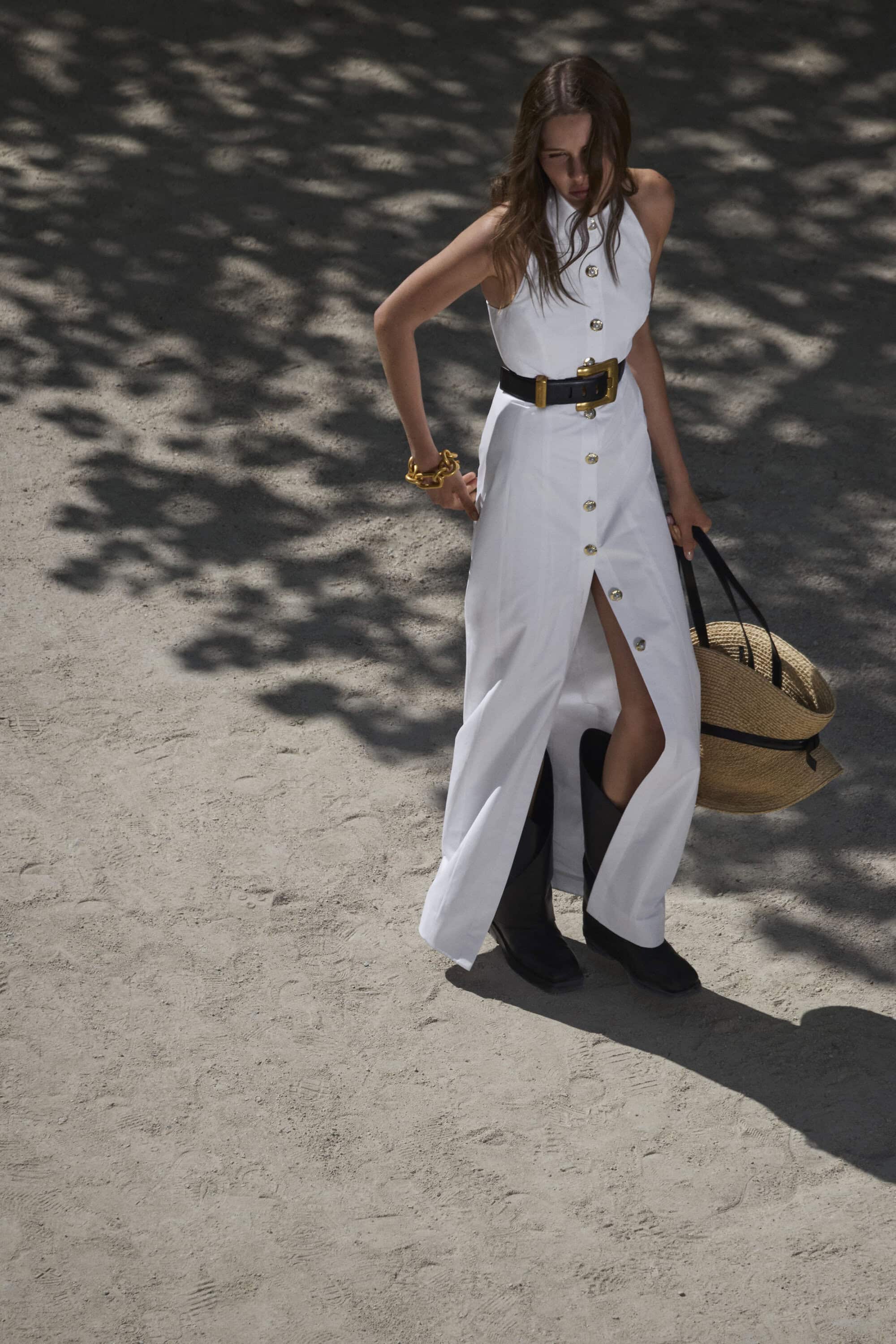 Model in a white buttoned maxi dress and black boots holding a straw tote bag outdoors in dappled sunlight.
