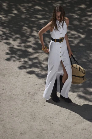 Model in a white buttoned maxi dress and black boots holding a straw tote bag outdoors in dappled sunlight.
