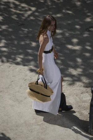 Model in white halter dress wearing a black belt and carrying a large straw tote bag walking outdoors