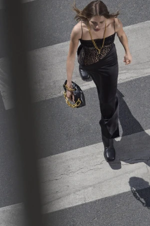 Model in black dress walking across a zebra crossing carrying a black handbag with a chunky gold chain