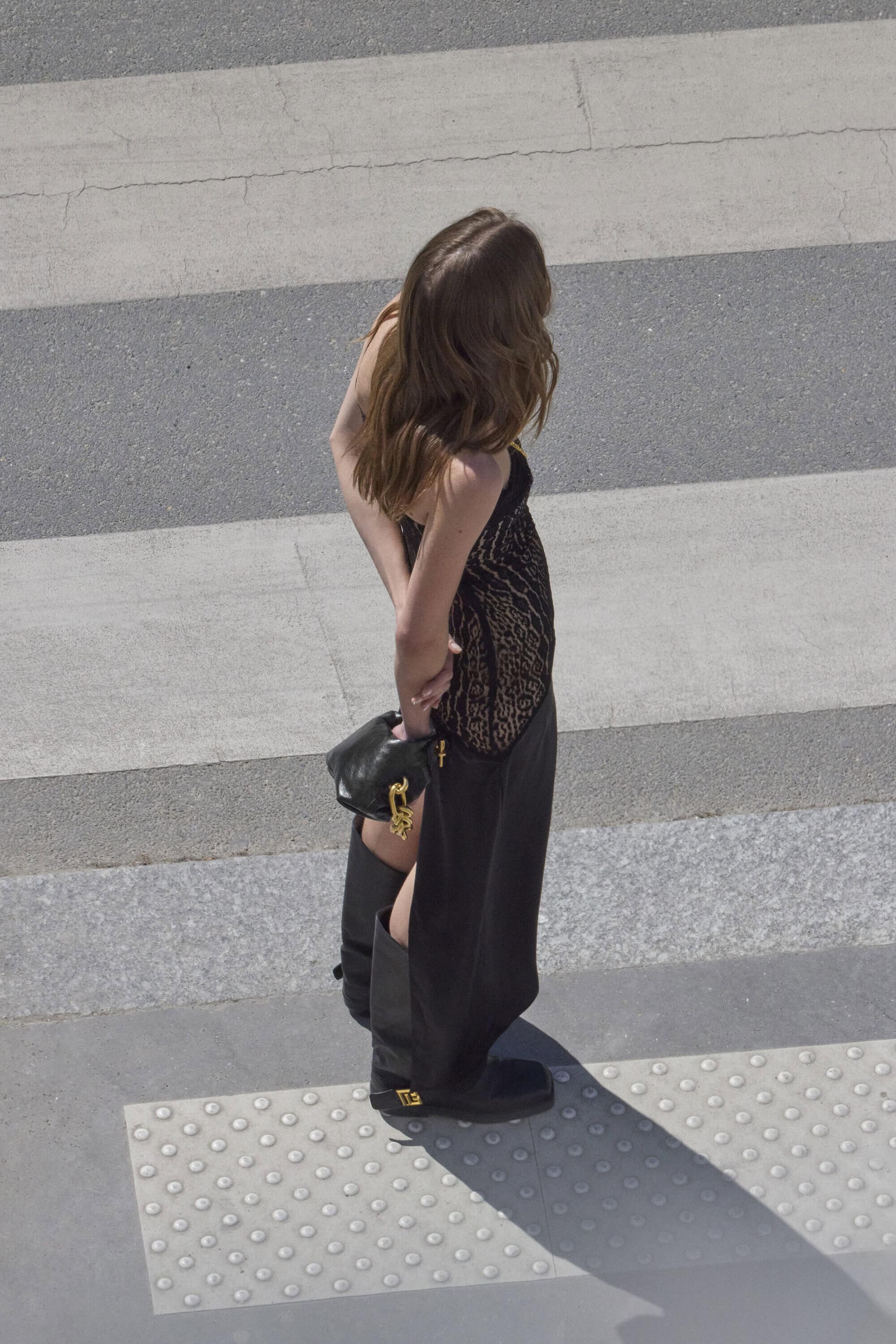 Model standing on a paved street crossing wearing a black dress and boots holding a handbag.