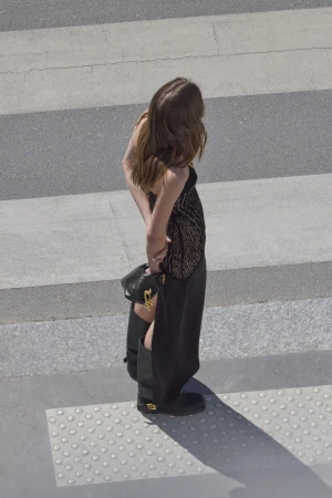 Model standing on a paved street crossing wearing a black dress and boots holding a handbag.