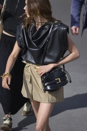 Model wearing a black leather top and khaki skirt holding a black handbag walking outdoors for Balmain pre-fall 2025 campaign