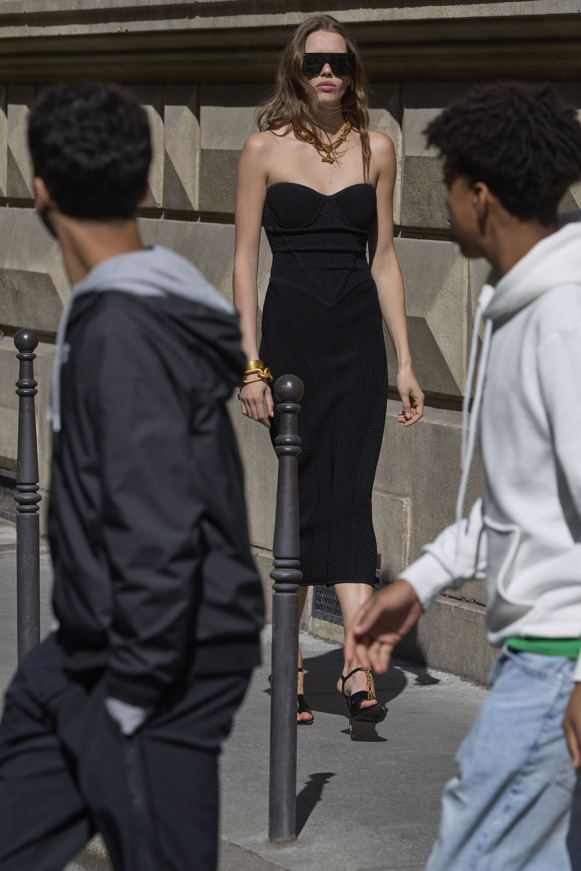 Model in a black dress and oversized sunglasses walking down a city street past two foreground figures