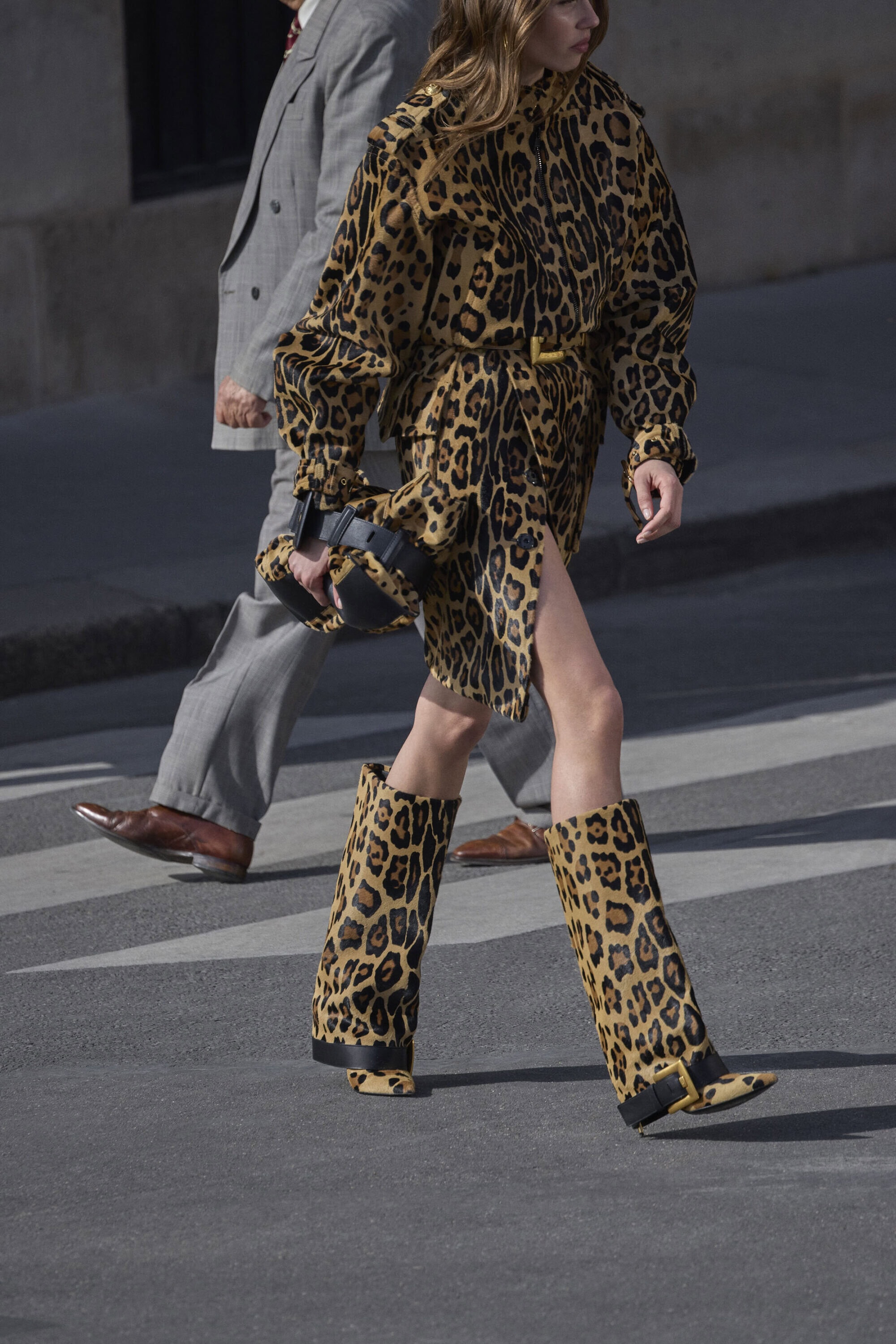 Model in a leopard print outfit and matching boots walking on a street next to a man in a grey suit