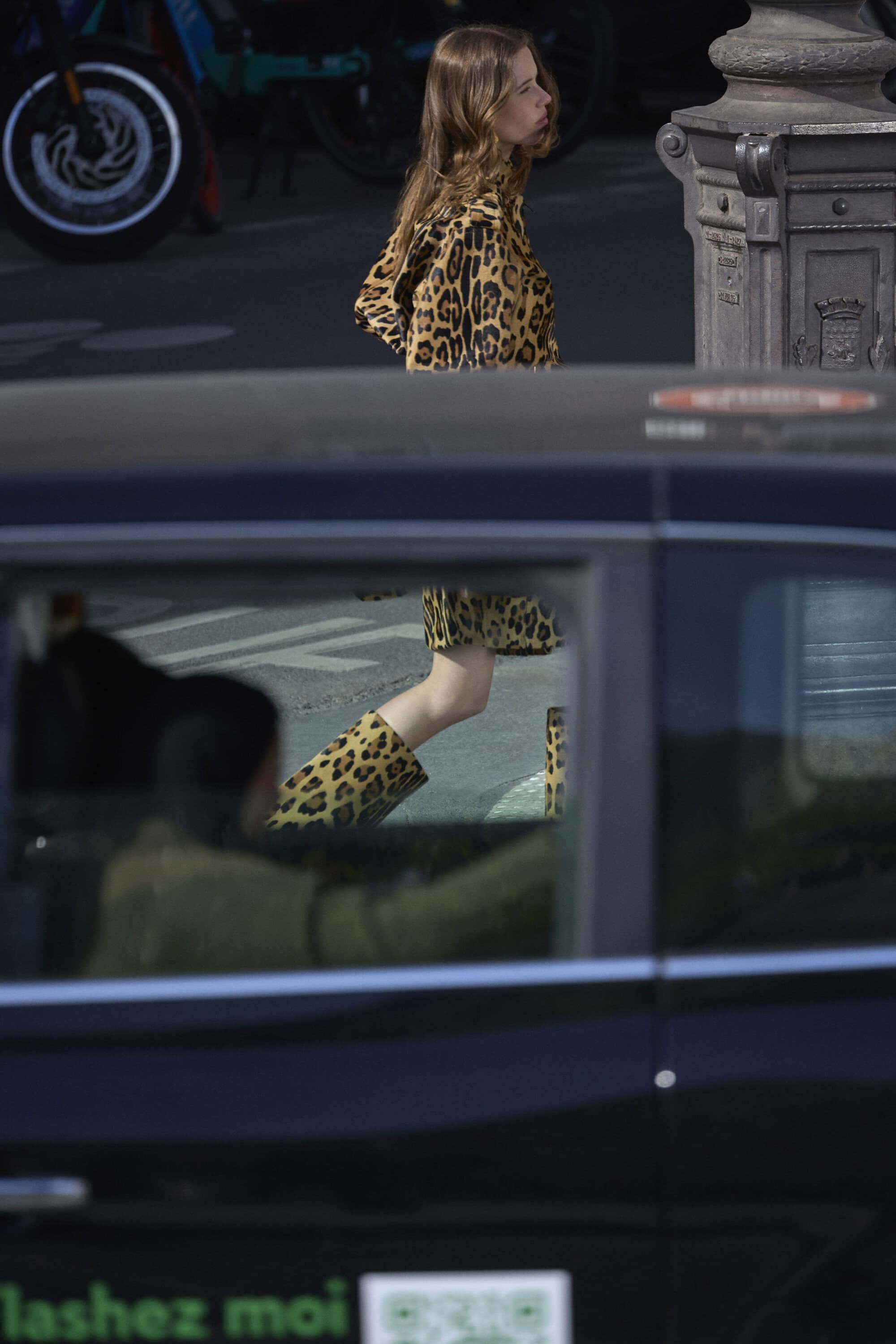 Woman in leopard print coat and boots walking past a car window in an urban street setting.