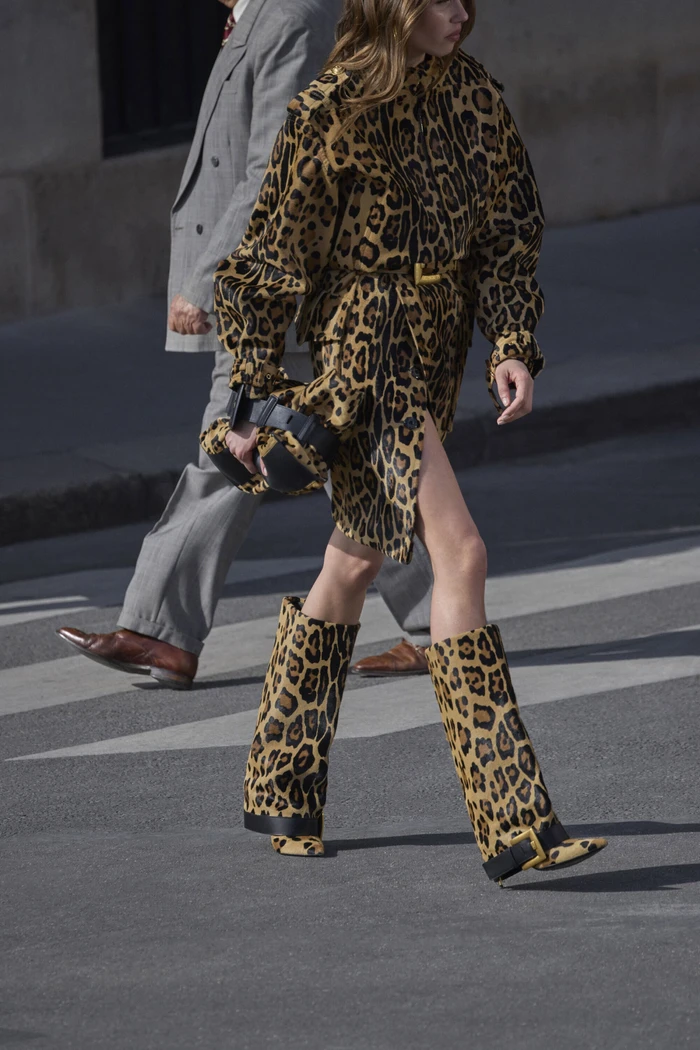 Model in a leopard print outfit and matching boots walking on a street next to a man in a grey suit