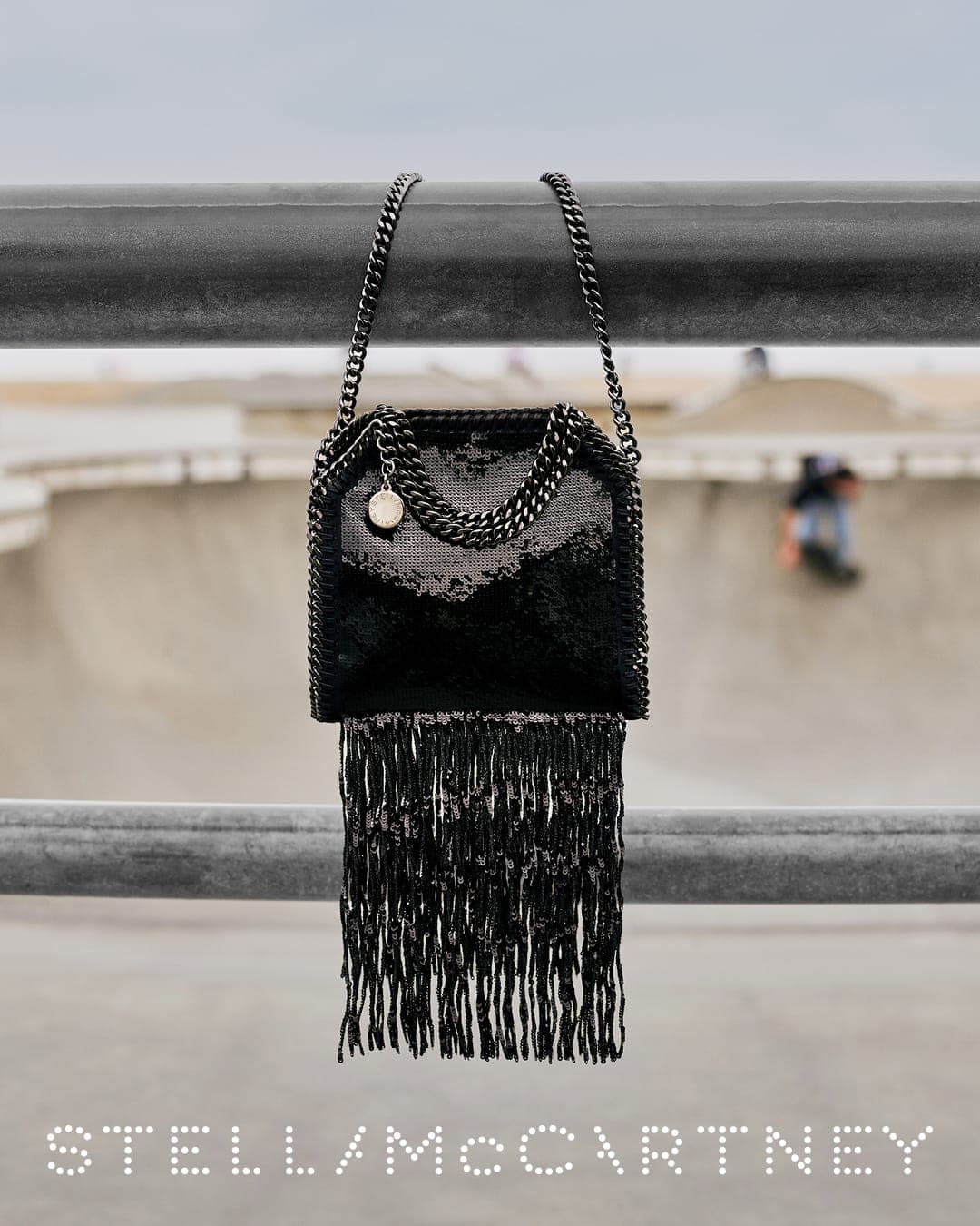 Black fringed chain handbag hanging over a metal rail with a blurred skatepark in the background for Stella McCartney