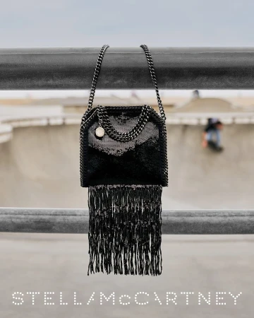 Black fringed chain handbag hanging over a metal rail with a blurred skatepark in the background for Stella McCartney