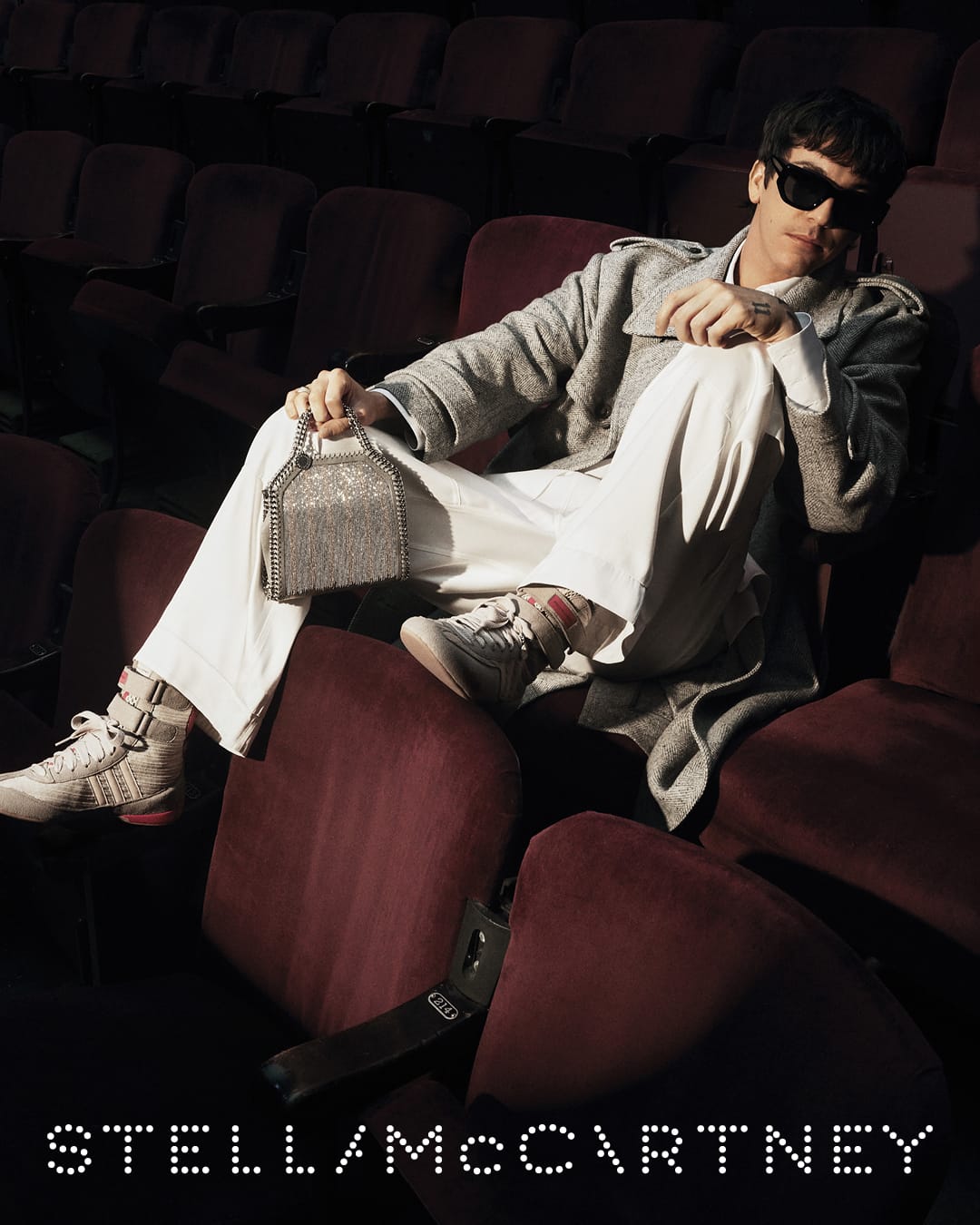 Model wearing a grey coat, white trousers, and sunglasses holding a metallic handbag while seated in a theater.