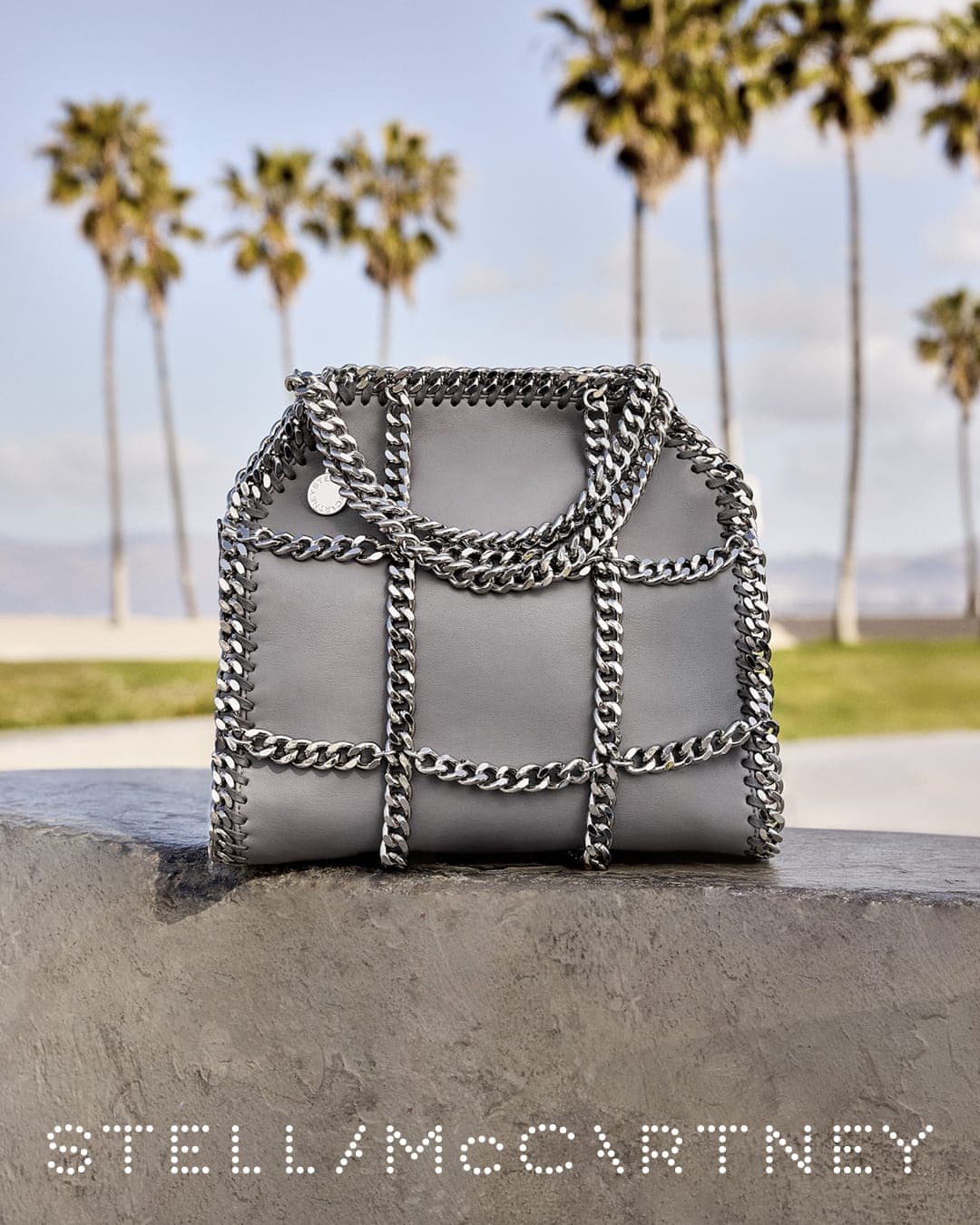 Grey handbag with heavy silver chain trim resting on a textured ledge with palm trees under an overcast sky.