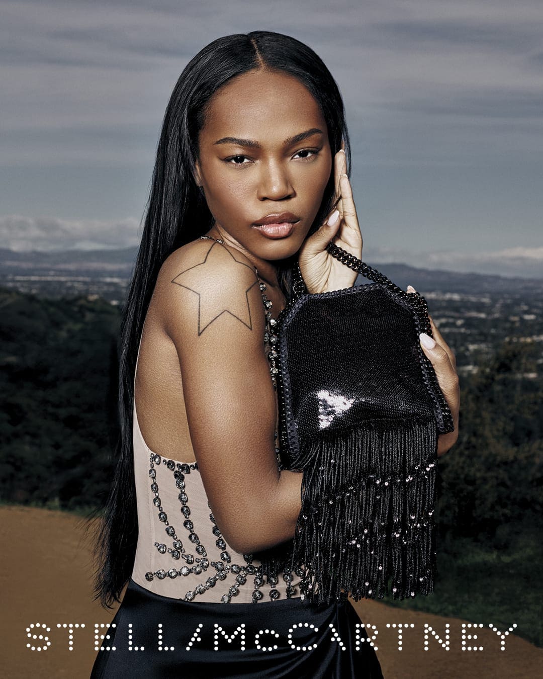 Model holding a black fringed handbag with a star tattoo on her shoulder against an outdoor landscape background