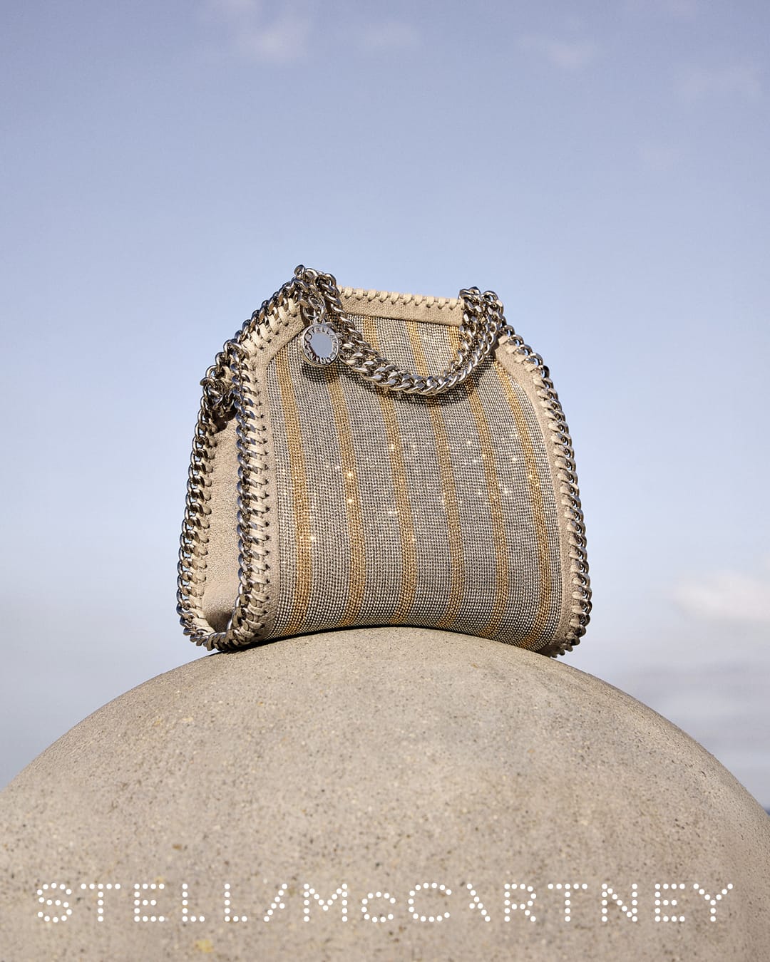 Stella McCartney Falabella handbag with chain trim perched on a rounded concrete sphere against a clear sky