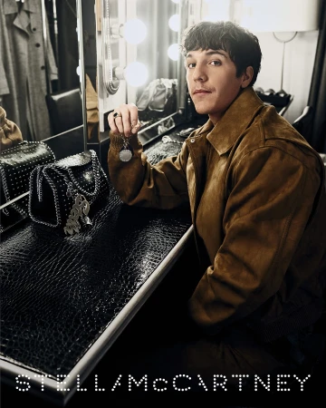 Man wearing a brown suede jacket seated backstage at a dressing table with black studded handbags for Stella McCartney campaign
