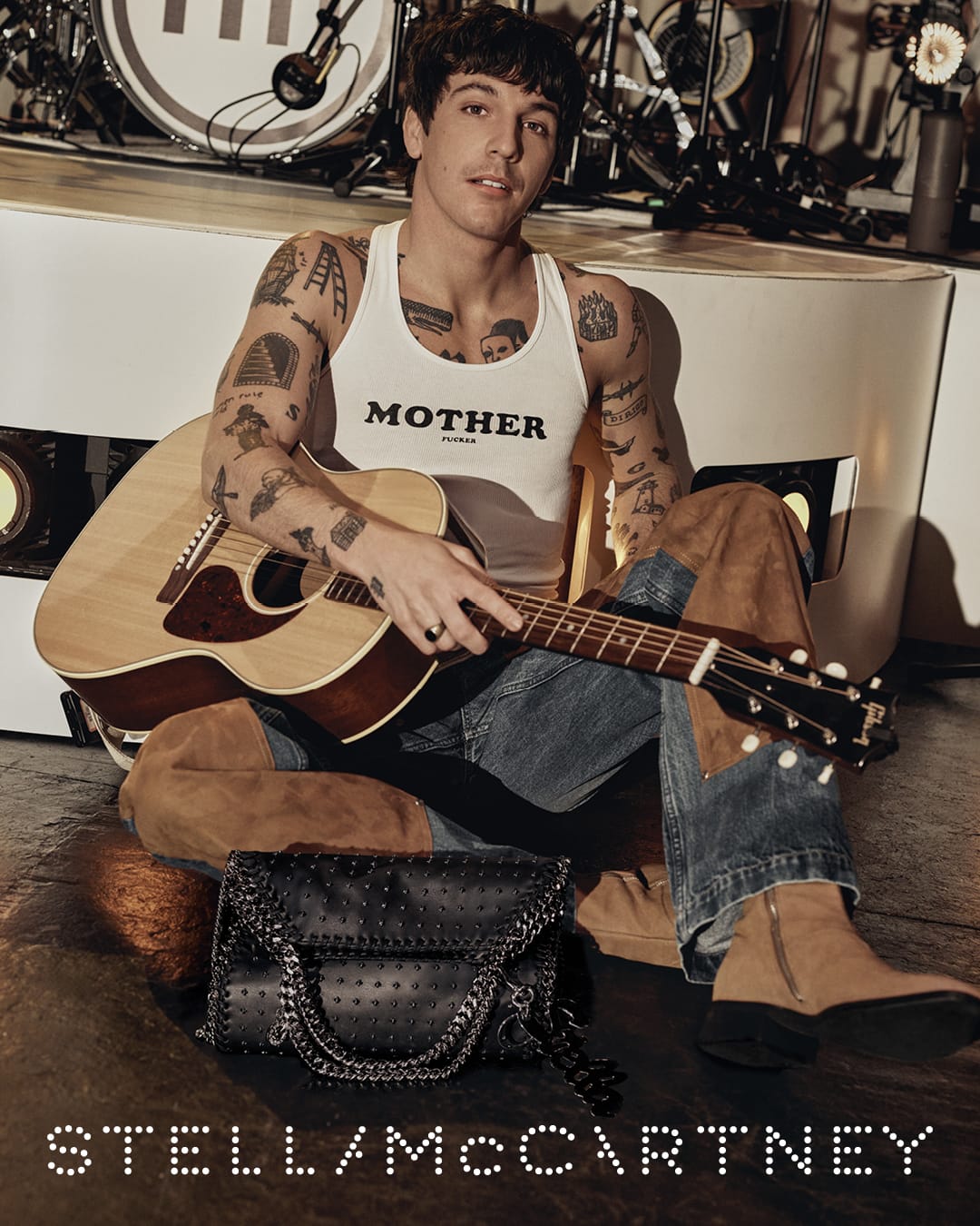 A tattooed man wearing a white tank top sits cross-legged on the floor playing an acoustic guitar with a black handbag nearby.