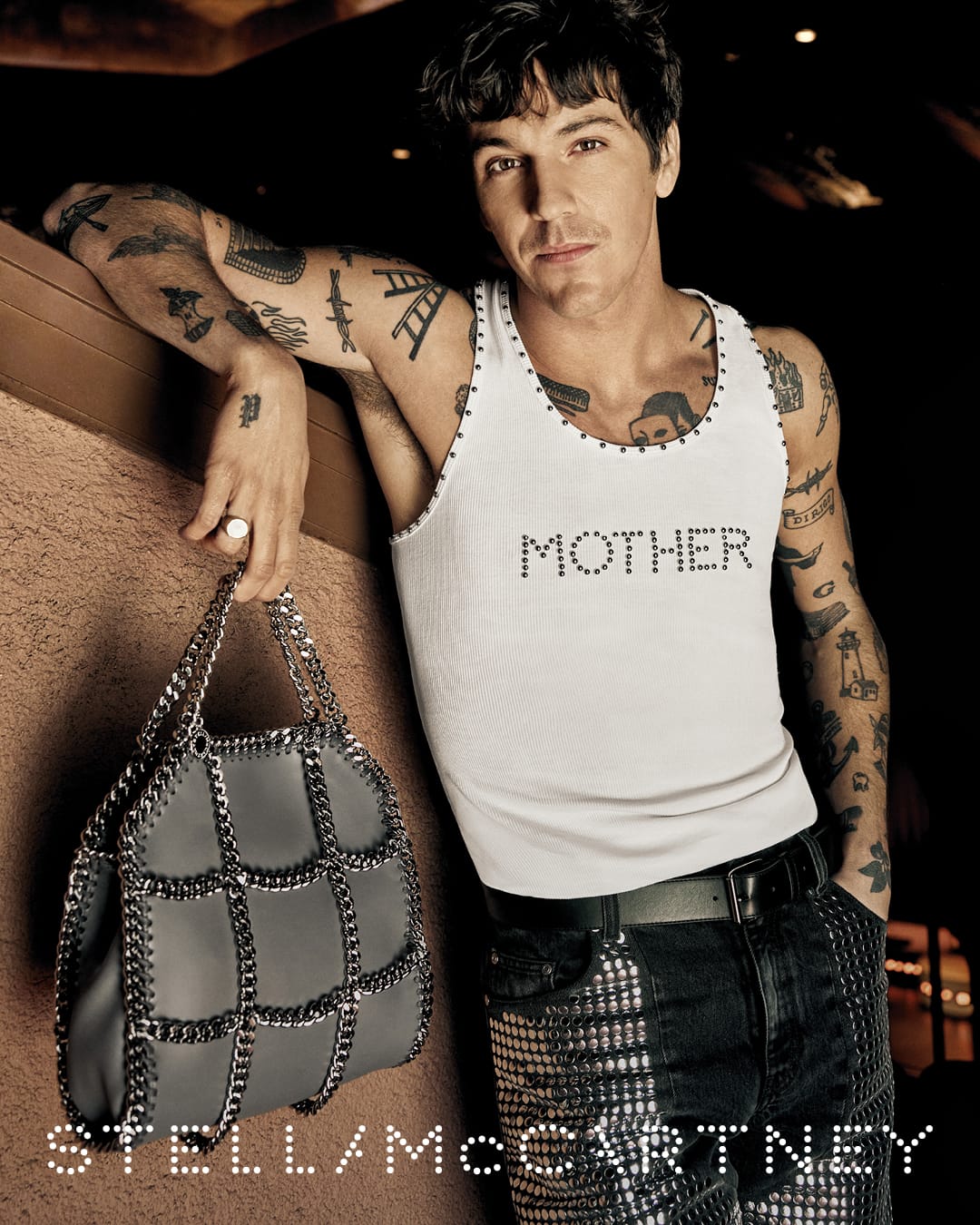 Tattooed man in a white studded tank top holding a grey chain-strap handbag for Stella McCartney