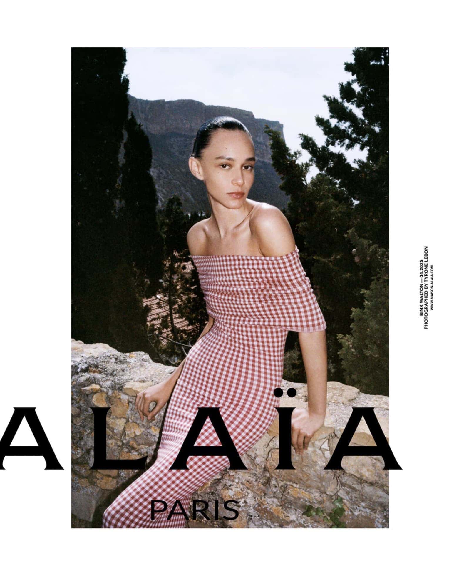 Model Binx Walton in a pink and white off-the-shoulder gingham dress leaning against a stone wall outdoors.