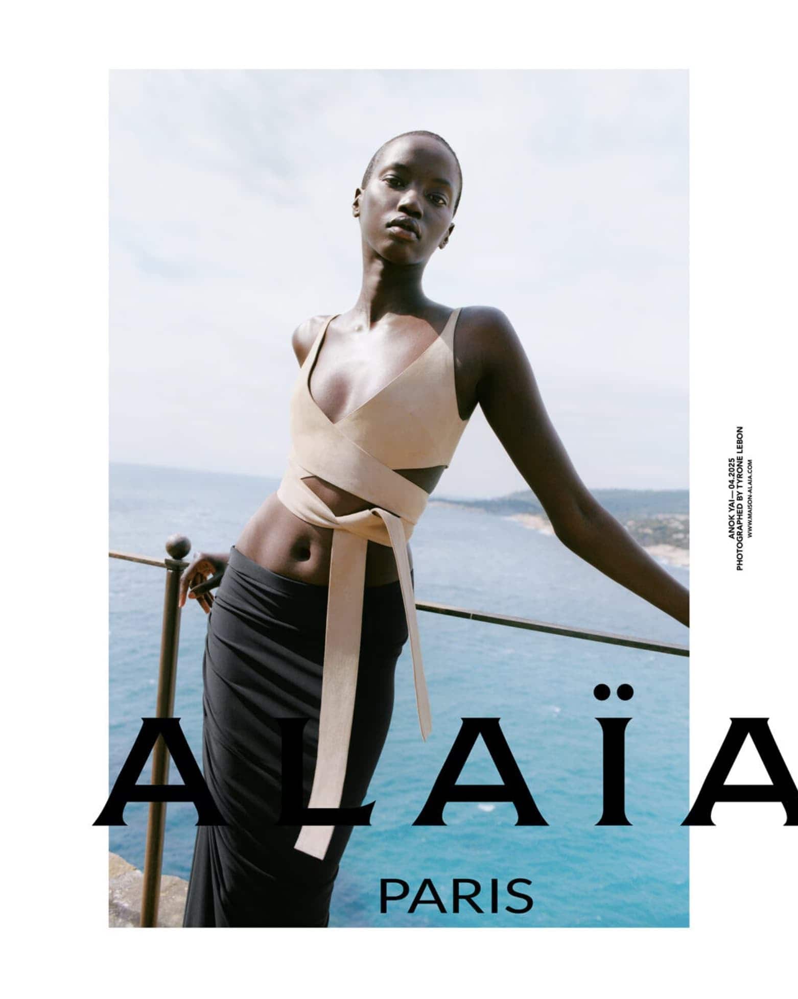 Anok Yai wearing an Alaia beige wrap crop top and black skirt against an ocean view