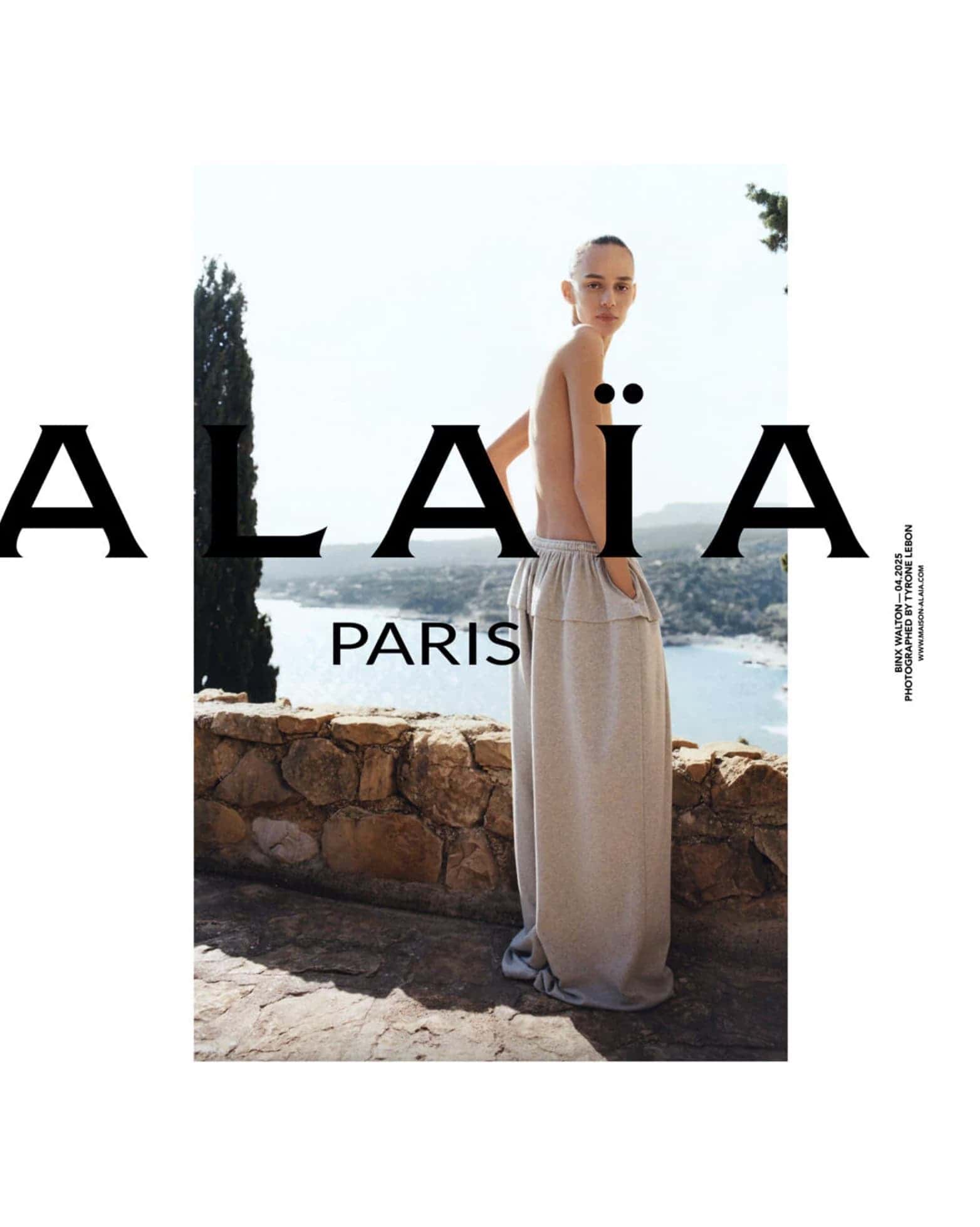 Model wearing loose trousers leaning against a stone wall overlooking water under a clear sky for an Alaia campaign.