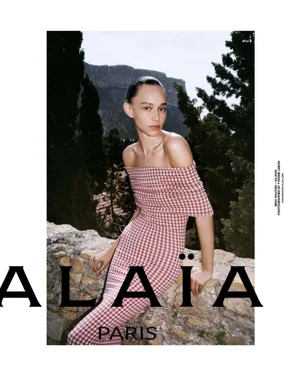 Model Binx Walton in a pink and white off-the-shoulder gingham dress leaning against a stone wall outdoors.