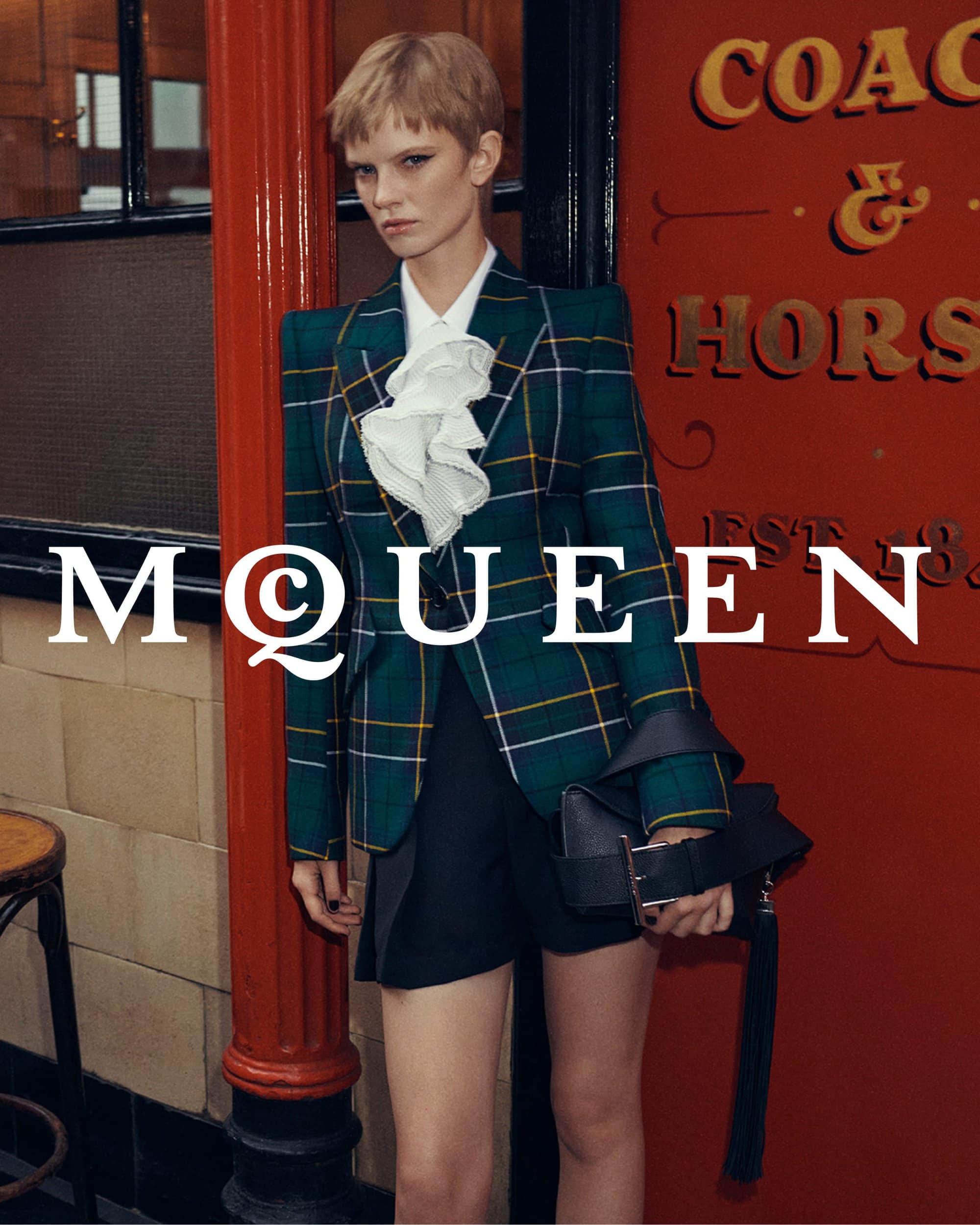 Model in a green plaid blazer and white ruffled shirt standing outside The Coach and Horses pub in London for McQueen.