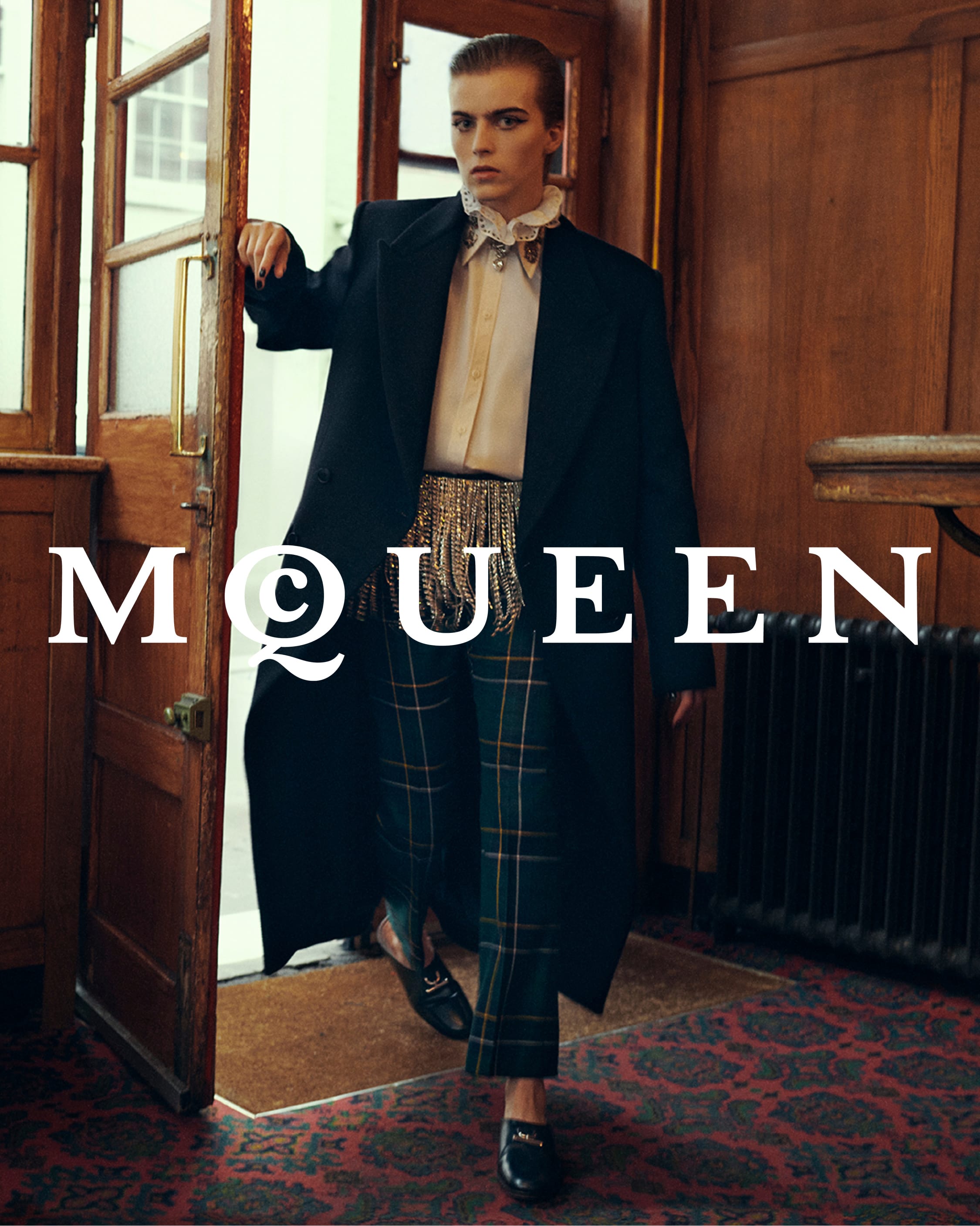 Model wearing a dark coat and plaid trousers standing in a wooden pub interior for McQueen Pre-Fall 2025 campaign.