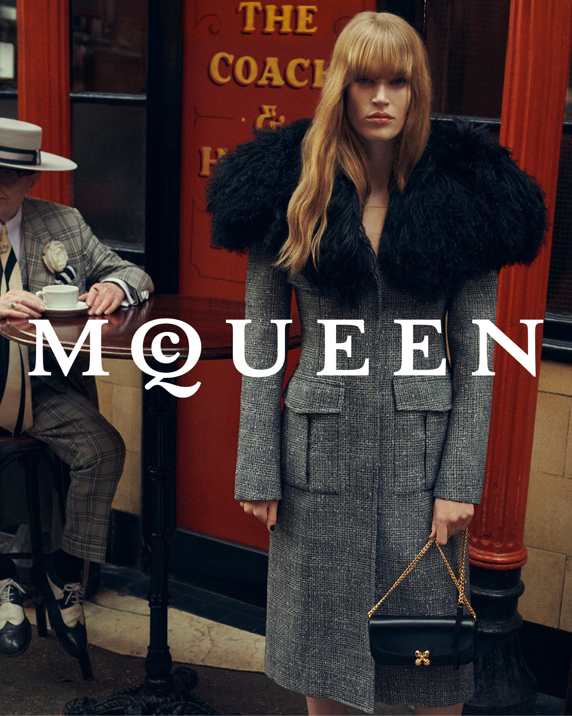 Model in a grey tweed coat with a large black fur collar holding a black handbag outside a pub