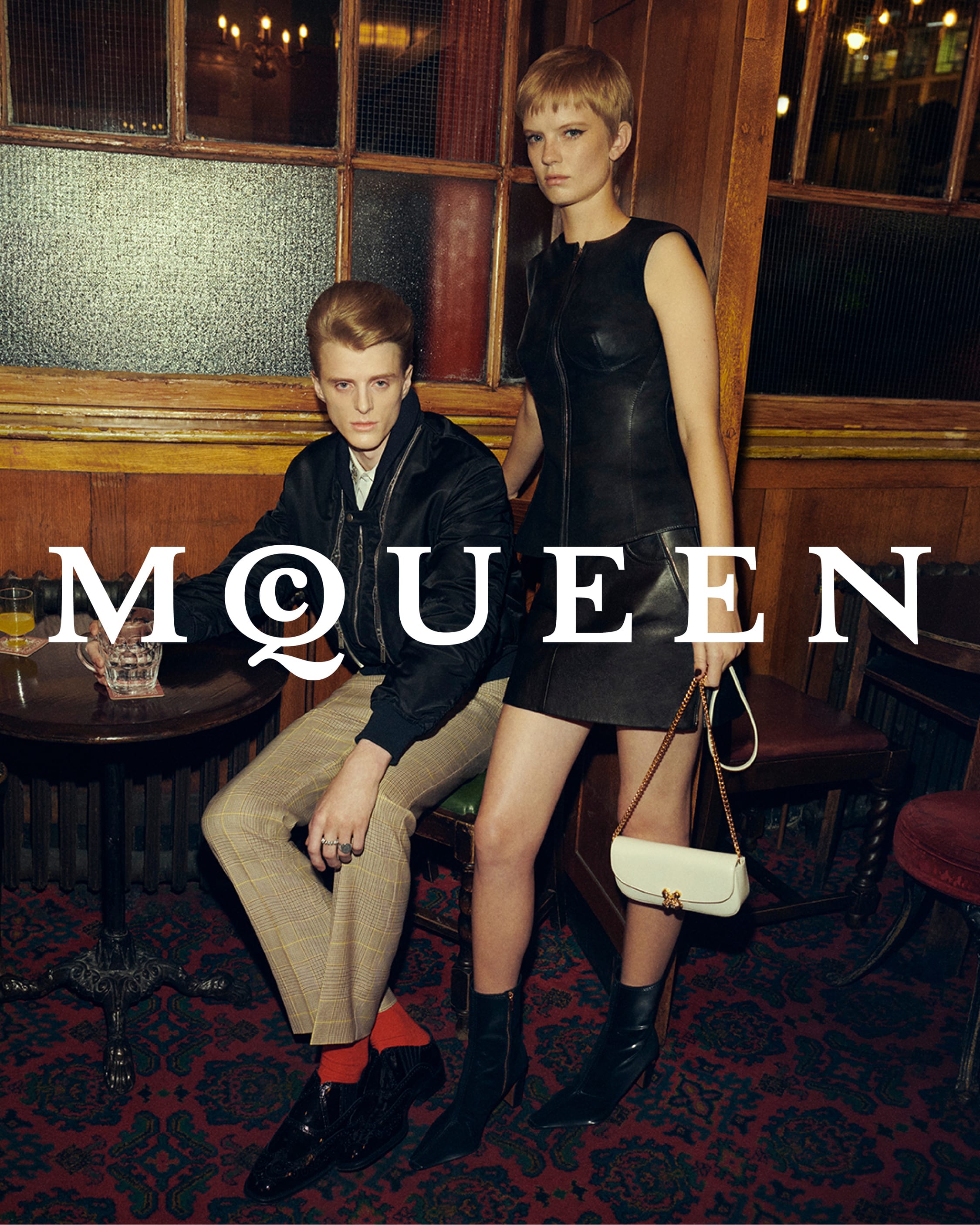 Two models inside a pub interior, a man seated in a dark jacket and trousers, and a woman standing in a black leather dress holding a white handbag