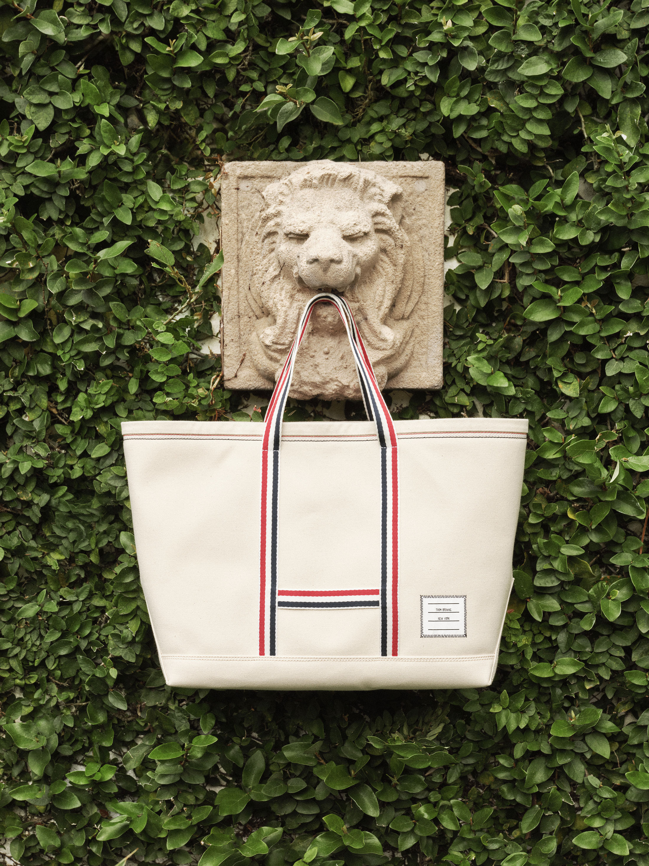 Off-white canvas tote bag with red, white, and blue straps hanging from a carved stone lion sculpture set against lush green ivy.