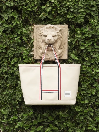 Off-white canvas tote bag with red, white, and blue straps hanging from a carved stone lion sculpture set against lush green ivy.