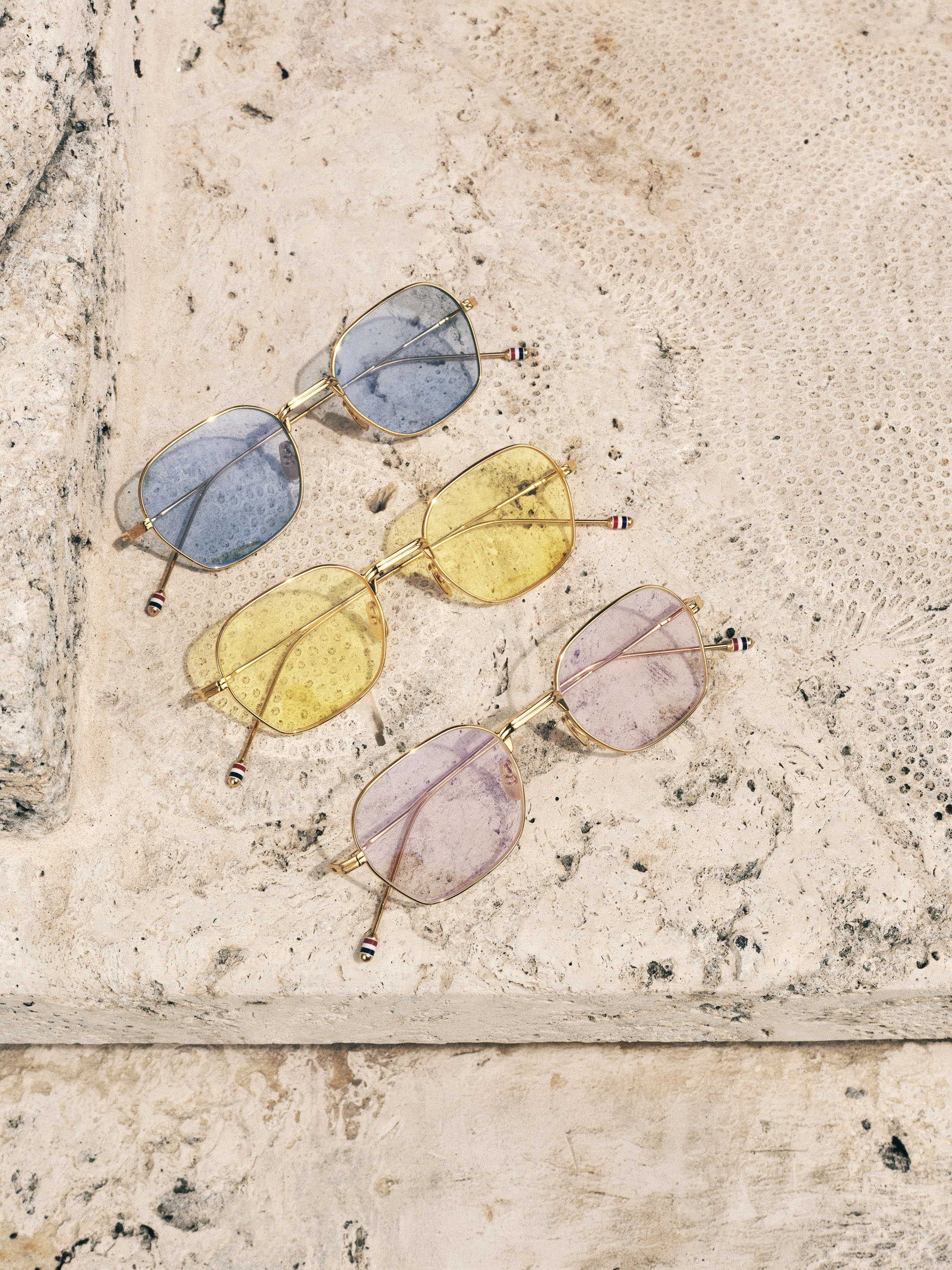 Three pairs of tinted designer sunglasses with gold frames displayed diagonally on a weathered stone surface.