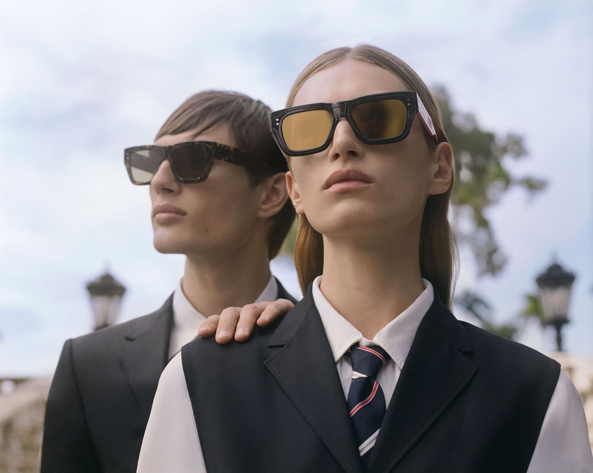 Two models wearing dark suits, white shirts, striped ties, and bold sunglasses against an overcast sky