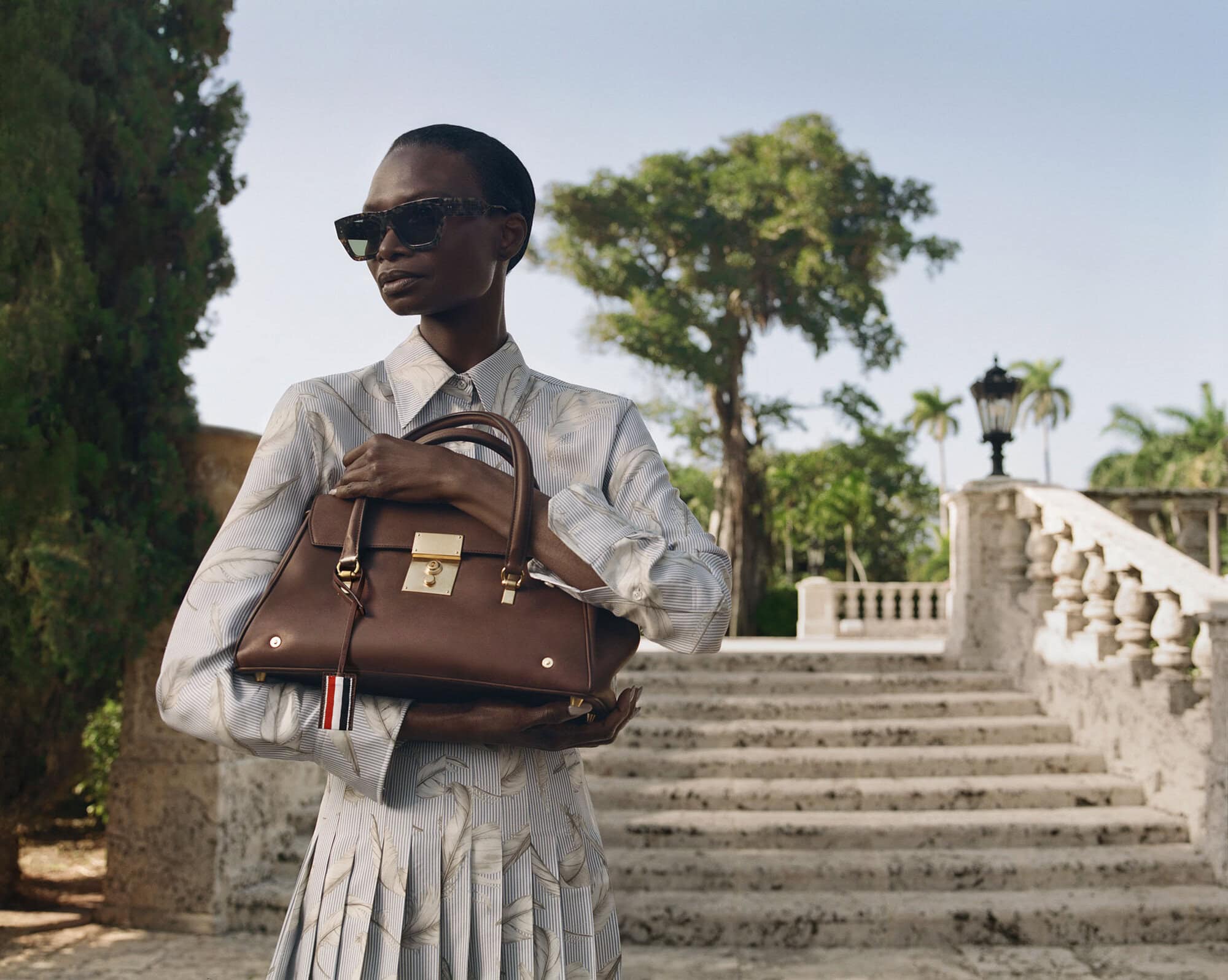 Model wearing dark sunglasses and patterned shirt holding a brown leather handbag outdoors near stone steps.