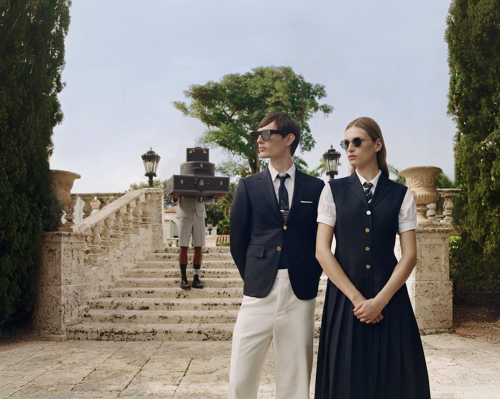 Two models wearing preppy navy blazers and sunglasses pose in front of stone outdoor stairs, with another person carrying luggage in background