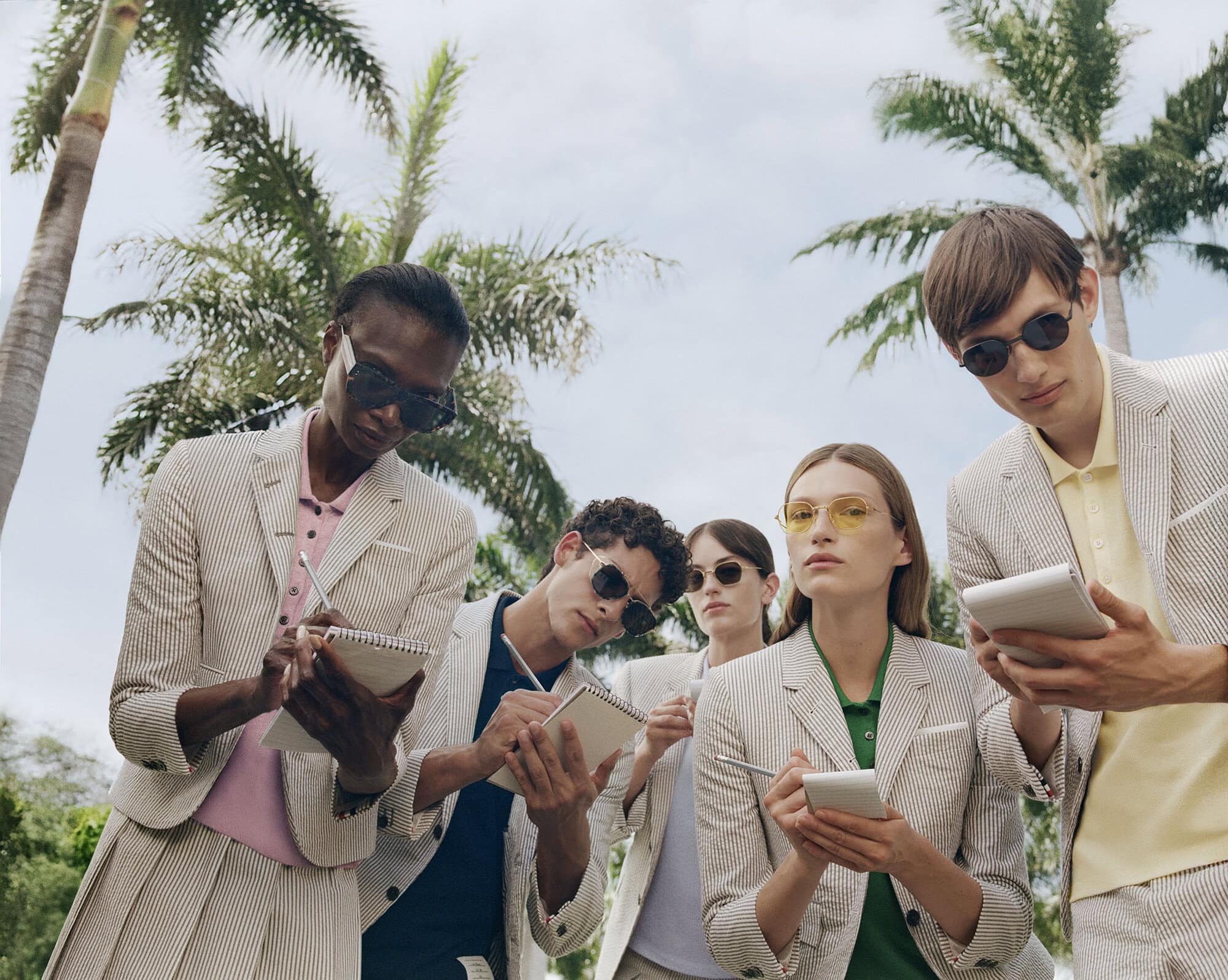 Group of models in striped blazers wearing sunglasses writing in notebooks outdoors with palm trees under an overcast sky.