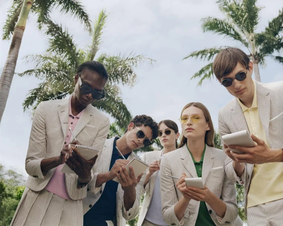 Group of models in striped blazers wearing sunglasses writing in notebooks outdoors with palm trees under an overcast sky.