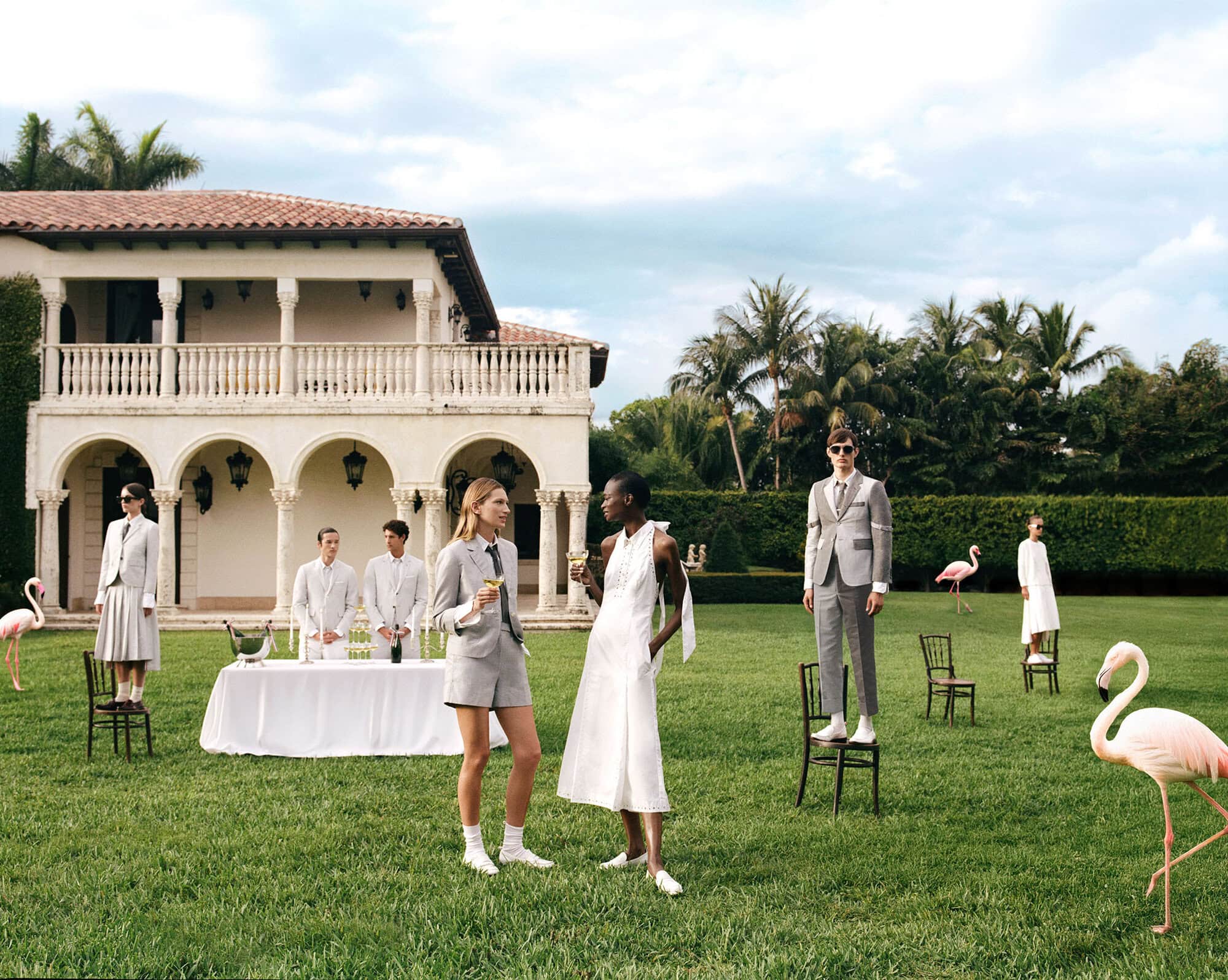 Group of models posing in a lush green lawn before a large white villa with pink flamingos standing around.