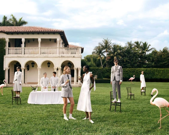 Group of models posing in a lush green lawn before a large white villa with pink flamingos standing around.