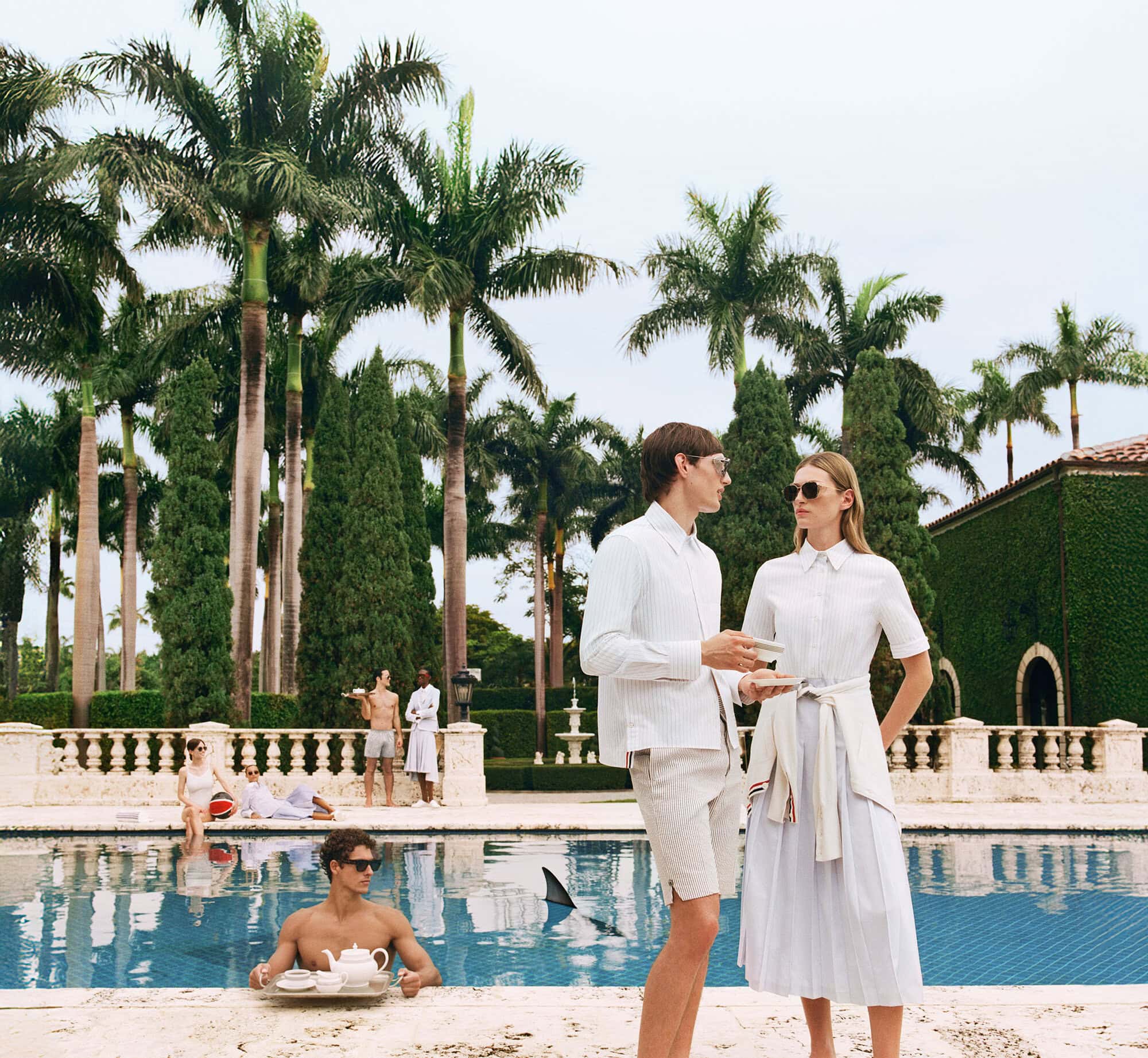 Models in white resort wear standing by a swimming pool surrounded by palm trees for Thom Browne Summer 2025 campaign.