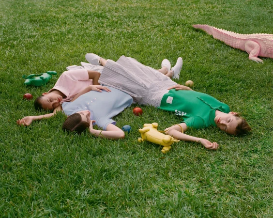 Three models lying on green grass wearing colorful polo shirts and skirts alongside novelty animal handbags